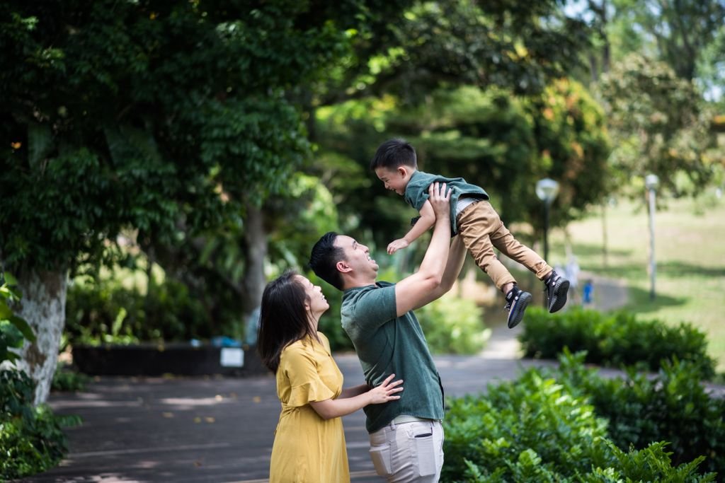 Family enjoying a playful moment in a vibrant green park. A father lifts his child as the mother watches with joy.